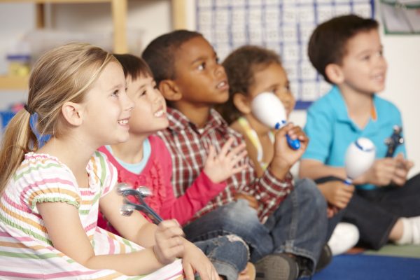 Five children sit alongside each other on the floor with their legs crossed, each holding a musical instrument.