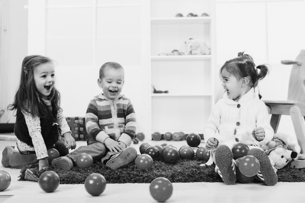 Black and white image of three children laughing together as they play with plastic balls.