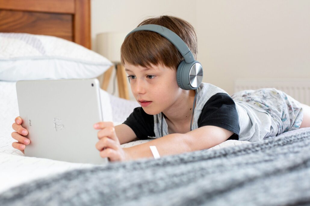 Young boy lying on a bed wearing headphones and using a tablet.